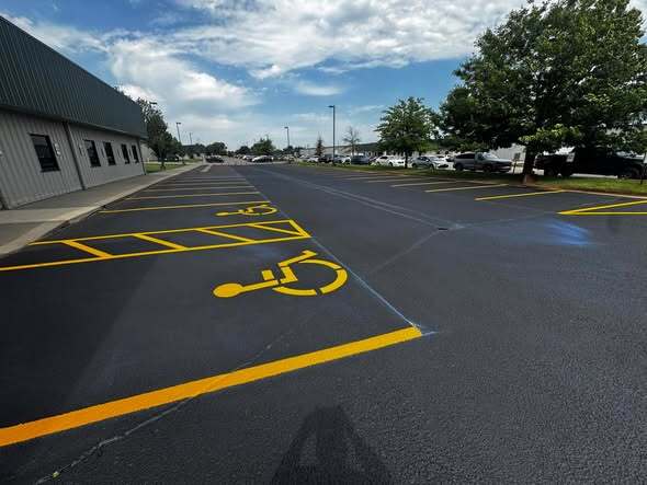 A freshly painted blue accessible parking symbol with white diagonal hatching in the access aisle beside it, next to a metal commercial building under a partly cloudy sky.