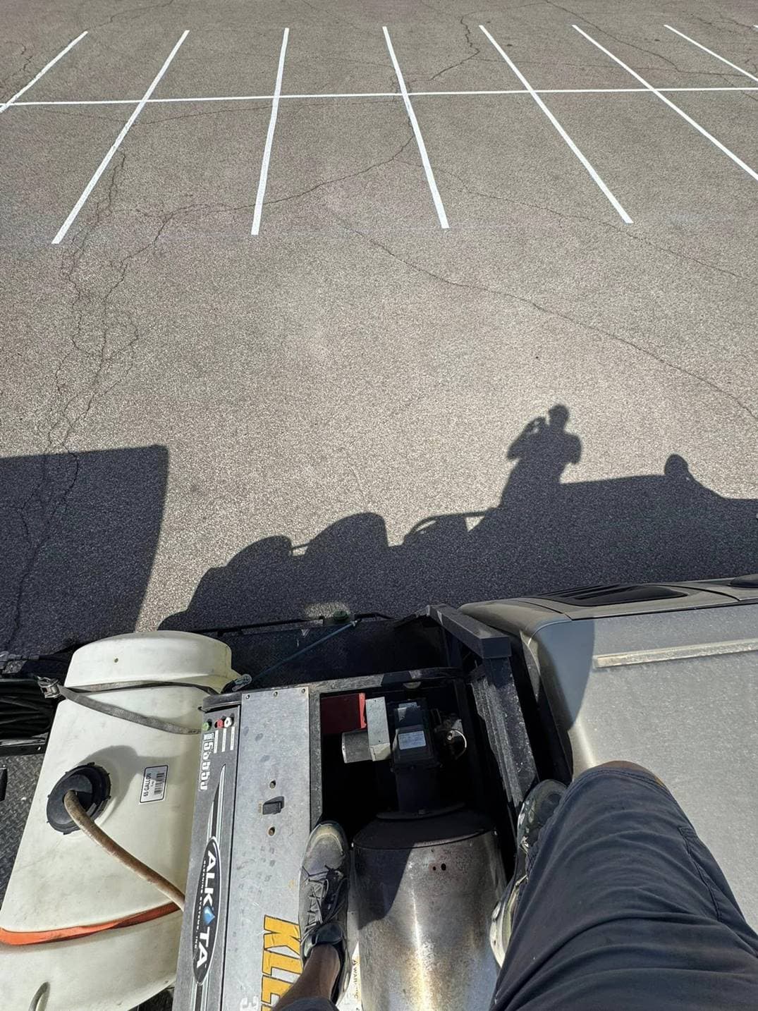 A point of view shot looking down at line striping equipment and a crew member's foot and long shadow cast across a freshly lined parking lot.