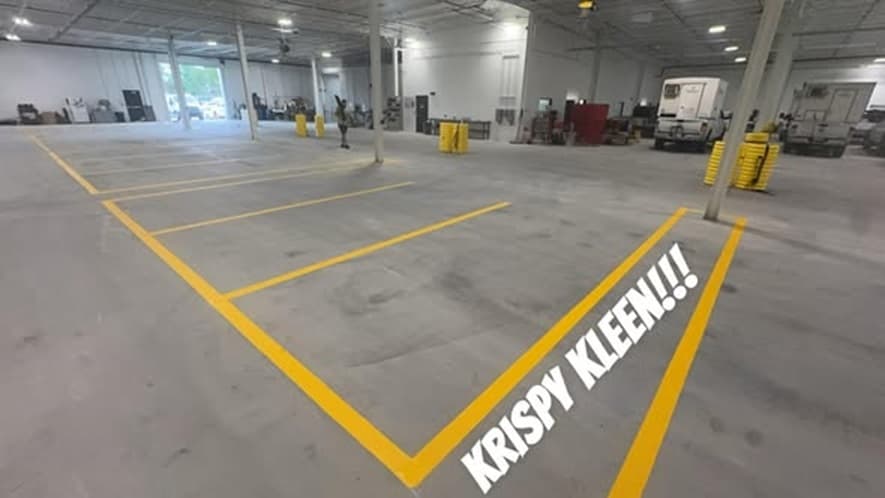 A yellow striped floor zone inside a warehouse, with a large custom text stencil painted across the concrete and a forklift and truck visible in the background.