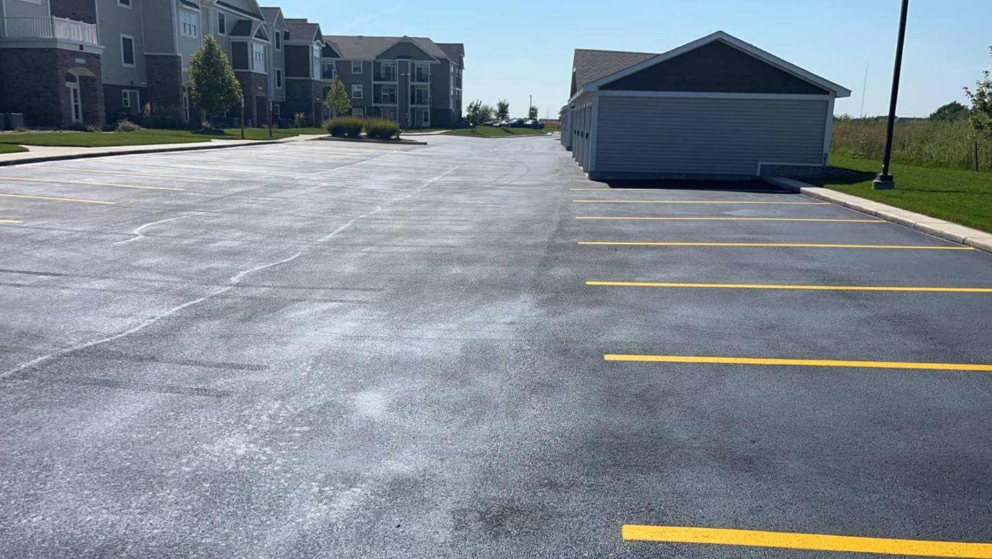 A freshly sealcoated apartment complex parking lot with new yellow stall lines, an attached garage building and residential units in the background.