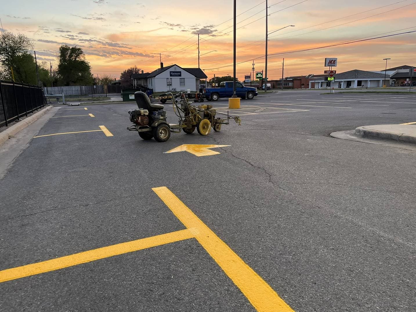 A ride-on line striping machine parked in a shopping center lot beside a freshly painted yellow directional arrow, photographed at dusk with storefronts lit in the background.