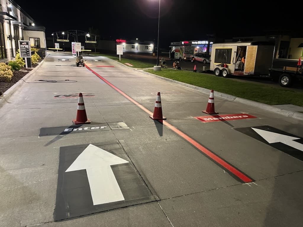 Custom directional lane stencils reading Enter and a lane marked for members, painted in white and red on pavement outside a business at night, with traffic cones set out.