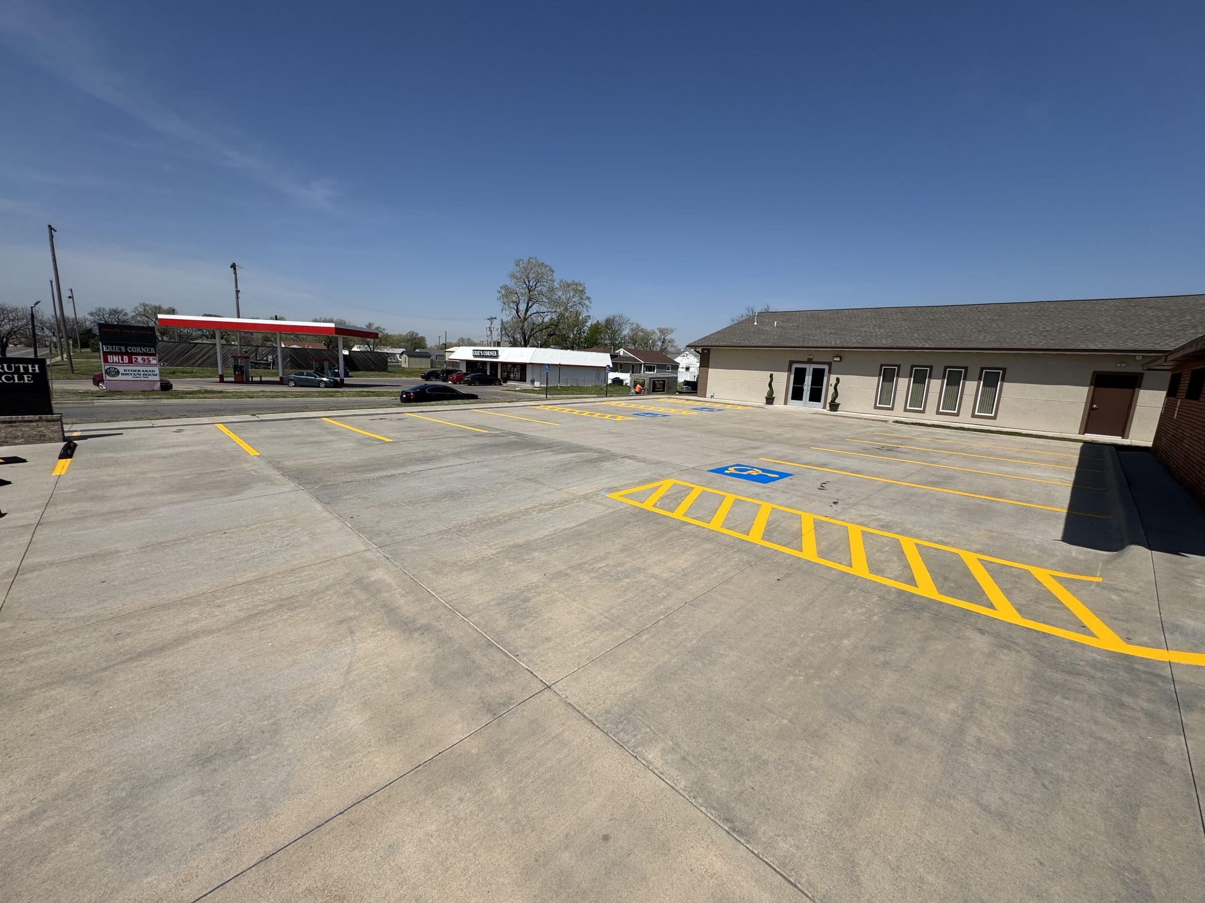 A concrete parking lot with new yellow stall lines, a long yellow hatched access aisle and a blue accessible stall symbol beside a single story building.
