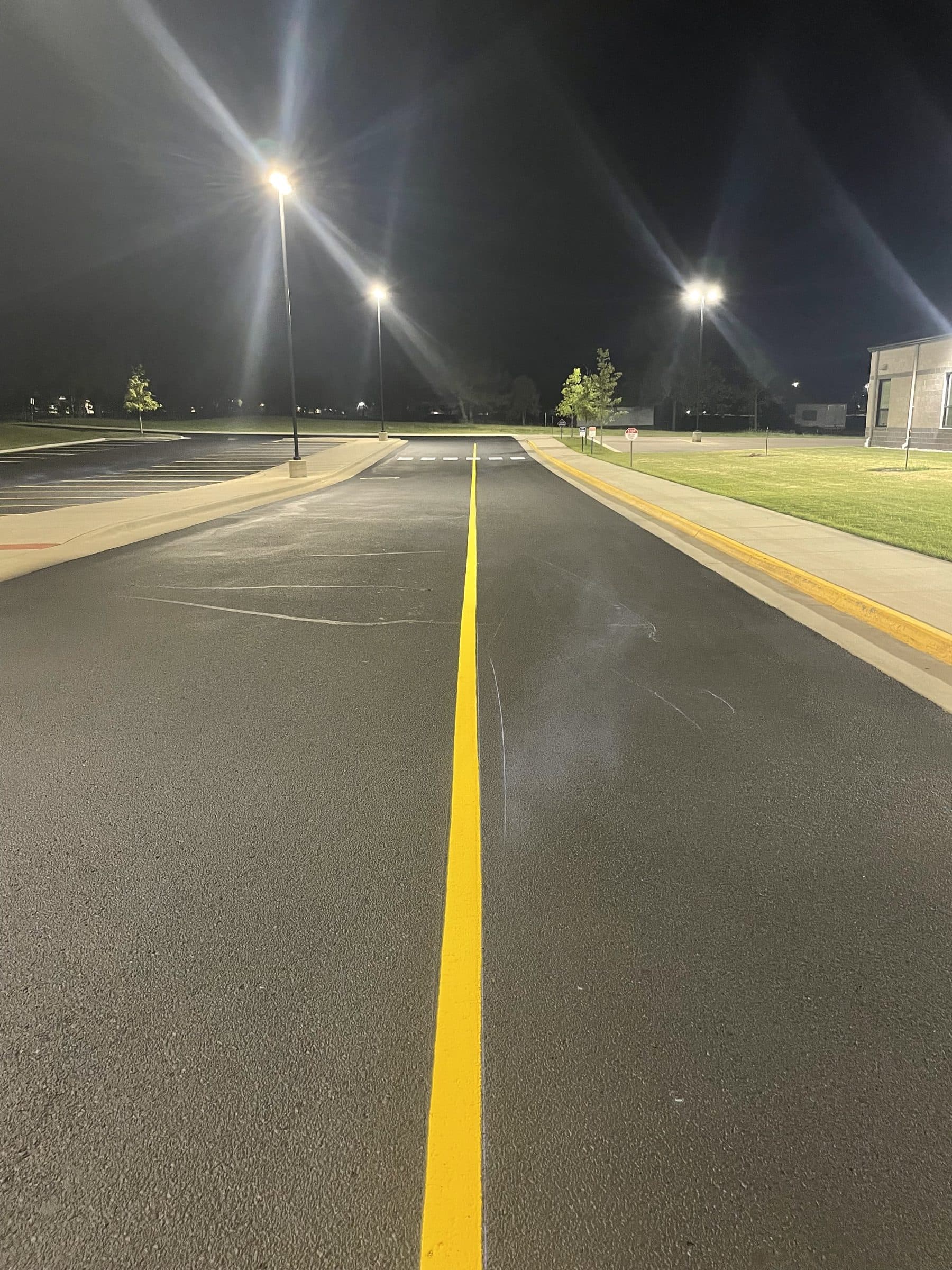 A fresh yellow center line running down a newly paved drive lane at night under tall parking lot lights, with a white stop bar at the far end.