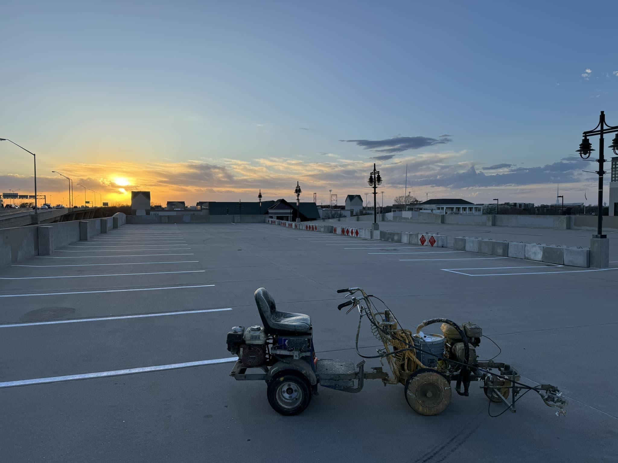A line striping machine parked on an upper deck of a parking structure at sunset, with fresh white stall lines and concrete traffic barriers marked with reflective diamonds.