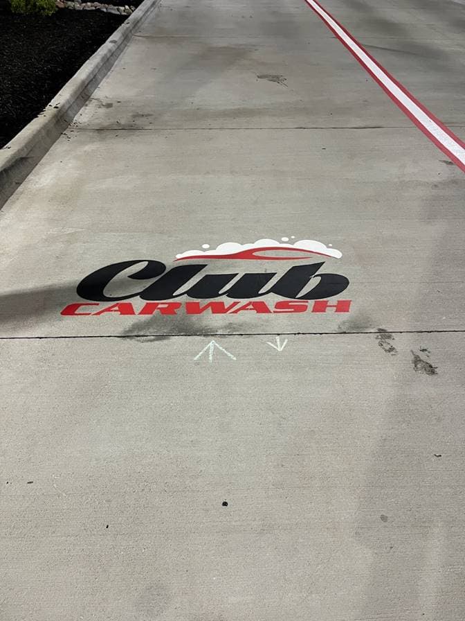 A custom logo stencil and directional arrows painted on a car wash drive lane, with a red guide line running alongside it.